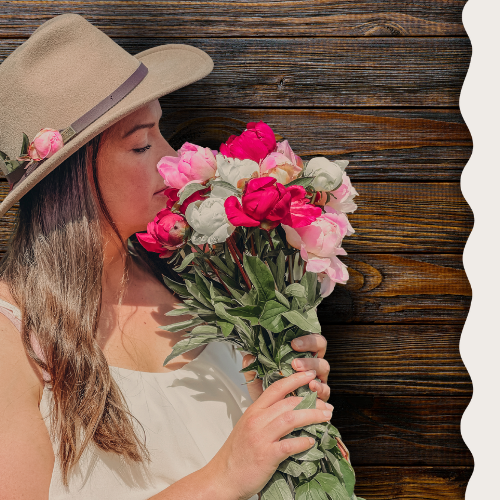 Woman wearing a hat with floral decorations holding a bouquet of flowers against a wooden background | Alaska Beauty Peony Cooperative | Homer, AK